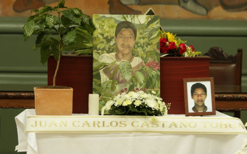 Parada junto al cofre donde reposa su hermano, quien estuvo desaparecido durante 23 años, Martha Isabel no deja de llorar. Cada tanto abre la caja de madera y toca las estructuras óseas que hay en el interior. Sus parientes se acercan y lloran junto a ella. Algunos la abrazan y otros miran la pintura de un cafetal en la que se ve a un muchacho moreno, de nariz achatada y cabello crespo recolectando granos de café. Ese joven es Juan Carlos Castaño Tobón, cuyo cuerpo fue entregado de manera digna por la Unidad de Búsqueda de Personas dadas por Desaparecidas (UBPD).