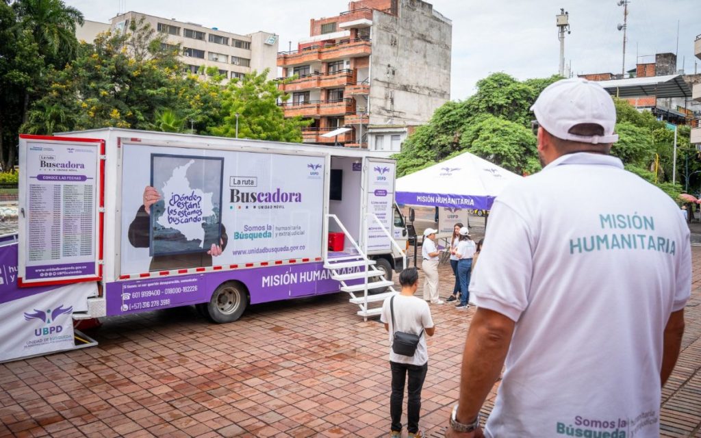 Hombre de espaldas vistiendo camiseta de la 'Misión Humanitaria' de la UBPD frente a la unidad móvil 'La ruta Buscadora'.