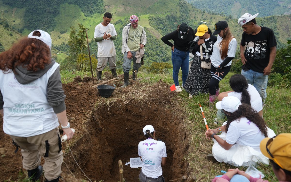 Vista superior desde el borde de una fosa recién excavada. En el fondo del hueco se encuentra un integrante del equipo forense con uniforme de la UBPD anotando datos. Alrededor de la fosa, varios campesinos, familiares e integrantes de la misión humanitaria observan en silencio el procedimiento.