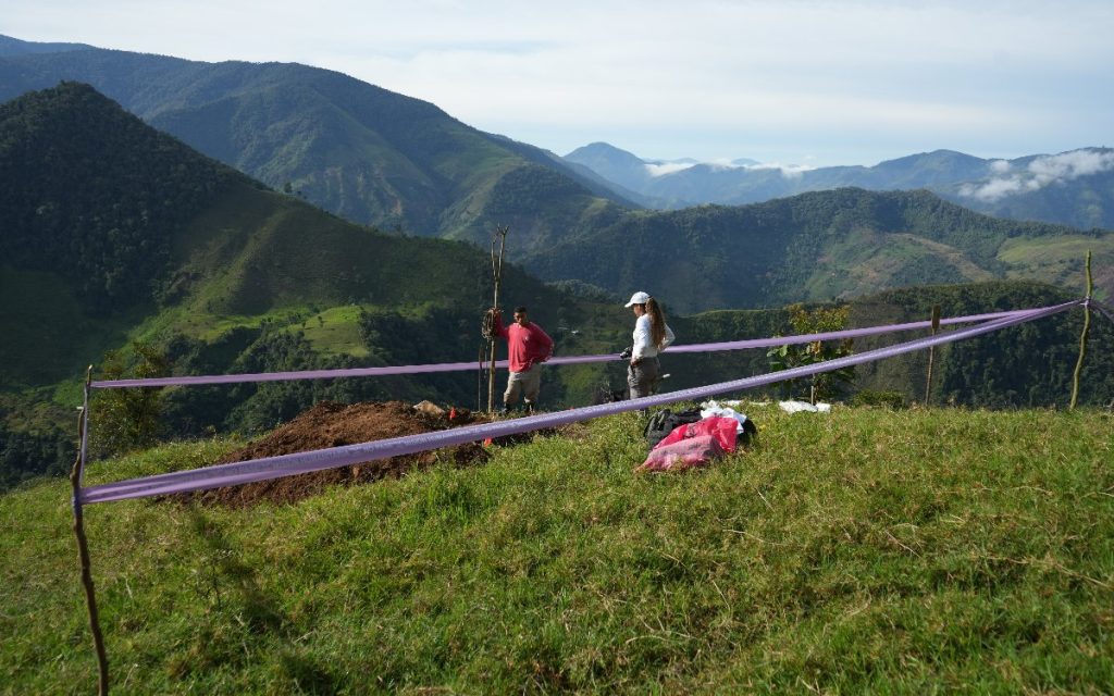 Dos personas se encuentran dentro de un perímetro acordonado con cinta morada de la UBPD sobre una cima de hierba verde. De fondo se aprecia un amplio valle rodeado por imponentes montañas e hileras de nubes. En el suelo hay bolsas con insumos de trabajo.