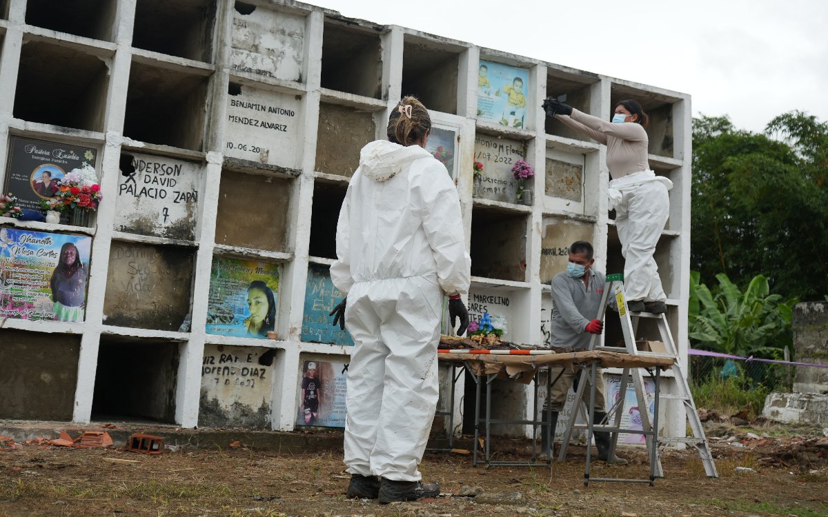 Personal con traje bioseguridad y tapabocas realizando labores de exhumación e inspección fotográfica frente a un bloque de bóvedas de cementerio.