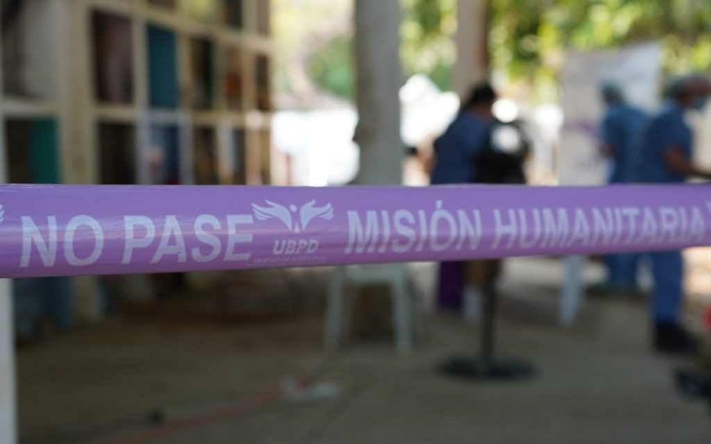 Primer plano de una cinta de seguridad de color lila que cruza horizontalmente la imagen con la inscripción 'NO PASE' y 'MISIÓN HUMANITARIA' junto al logotipo de la UBPD (Unidad de Búsqueda de Personas dadas por Desaparecidas). Al fondo, de manera difuminada, se observa a personal con trajes médicos de protección azul en un área exterior.