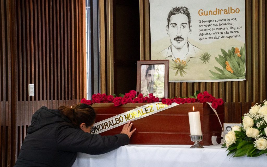 Una mujer inclina la cabeza y apoya la mano sobre el ataúd de Gundiralbo Morales, decorado con rosas rojas, un cirio y una cartelera con su rostro e historia.