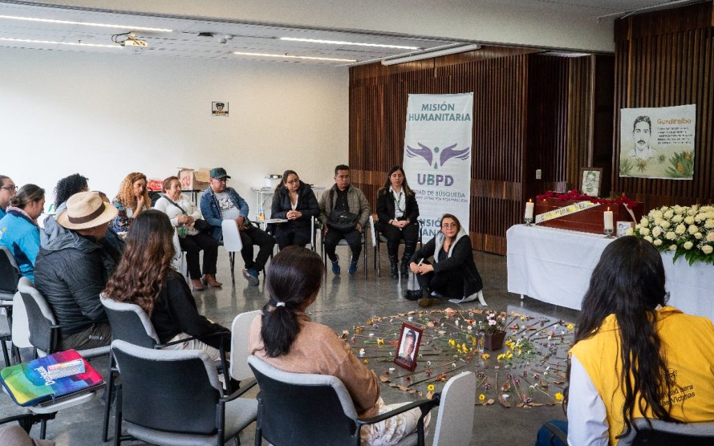 Personas e integrantes de la Unidad de Búsqueda de Personas dadas por Desaparecidas (UBPD) sentados en círculo alrededor de un altar con flores y una fotografía durante un acto ceremonial.