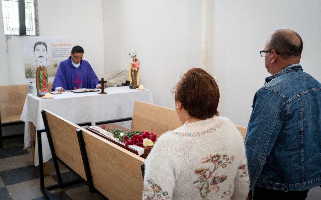 Un sacerdote frente a una mesa ceremonial oficia una misa frente a dos personas de espaldas y un féretro cubierto con ramos de flores rojas.