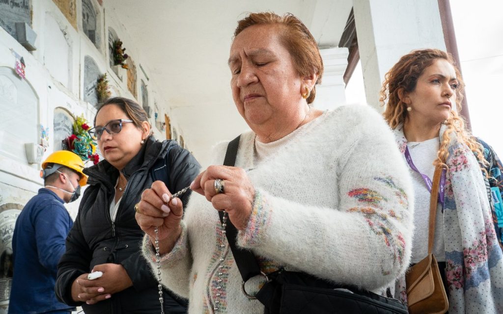 Una mujer con abrigo blanco sostiene atentamente un rosario entre sus manos con expresión reflexiva. A sus lados la acompañan dos mujeres en una galería de cementerio con bóvedas funerarias de fondo.