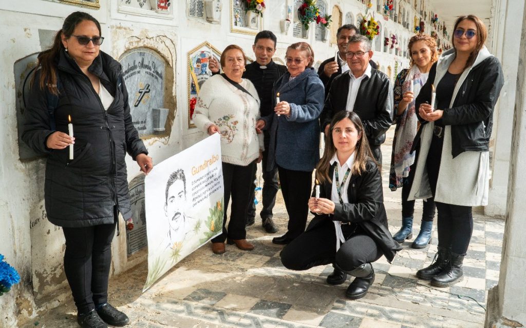 Un grupo de ocho personas reunidas en un pasillo de cementerio sostienen velas encendidas junto a la lápida grabada con el nombre "Gundiralbo Morales Dimate". En el frente sostienen un cartel con el dibujo de su rostro y un mensaje conmemorativo.