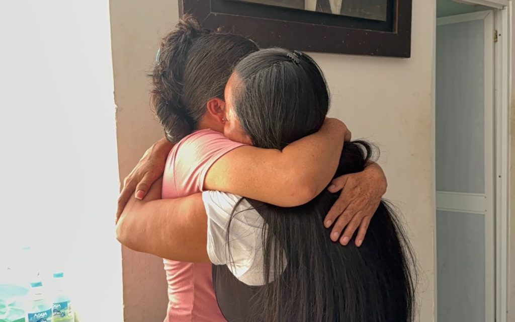 Dos mujeres se abrazan con fuerza. Una viste camiseta rosada y la otra una blusa blanca con chaleco o delantal oscuro y cabello largo. Sus brazos rodean los hombros y la espalda de la otra persona en una muestra de afecto emotiva.