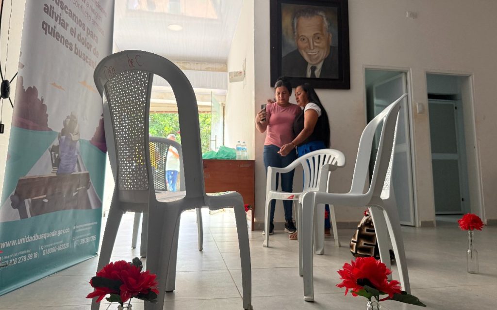 Vista amplia de un salón iluminado. Al fondo, dos mujeres permanecen abrazadas sosteniendo un teléfono celular frente a un cuadro en la pared. En el primer plano se observan sillas plásticas blancas con flores rojas decorativas a sus pies, y a la izquierda un pendón institucional de la Unidad de Búsqueda de Personas dadas por Desaparecidas.