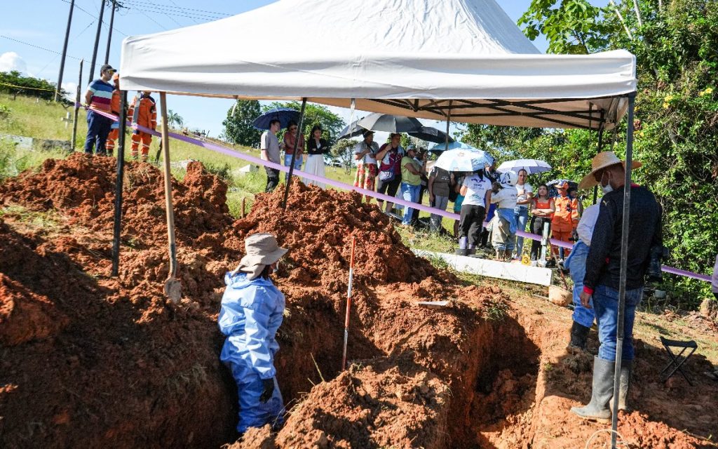 Un operario con traje de protección azul trabaja dentro de una zanja de excavación en un cementerio a campo abierto, protegido por una carpa blanca. Al fondo, tras una cinta delimitadora, varias personas observan el trabajo bajo parasoles.