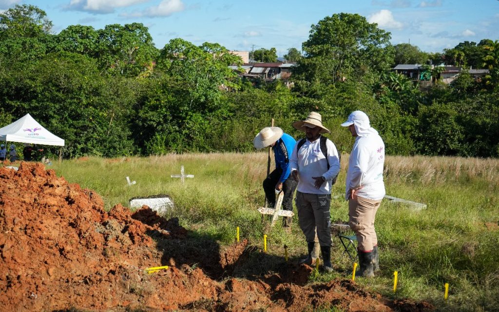 Tres integrantes del equipo de búsqueda de la UBPD conversan al borde de una fosa excavada en un terreno con cruces de madera. Al fondo se observa vegetación densa y casas del municipio.