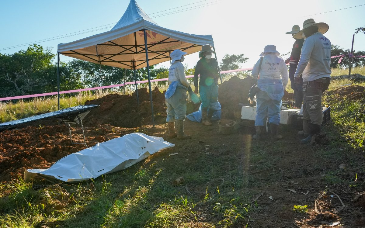 En primer plano, una bolsa blanca de recuperación de cuerpos descansa en el suelo cerca de una carpa. Alrededor, personal con trajes de bioseguridad y sombreros realiza labores técnico-forenses.