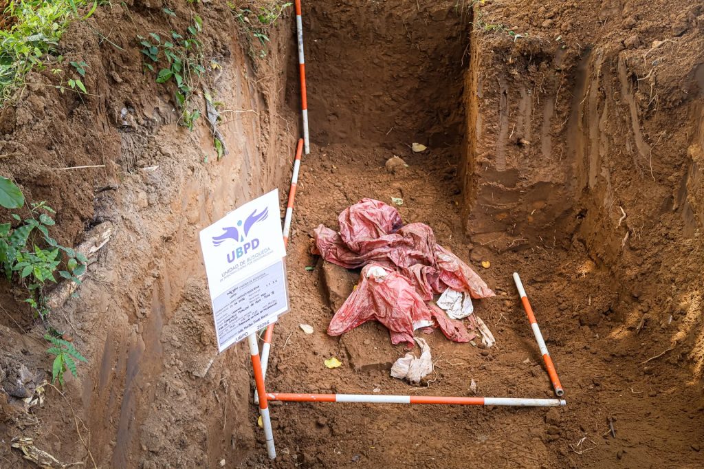 Vista superior de una fosa rectangular excavada en tierra marrón. En el centro hay elementos envueltos en plástico rojo demarcados por miras topográficas a rayas rojas y blancas. A la izquierda se observa una placa informativa con el logotipo de la UBPD y datos manuscritos del proceso de excavación.