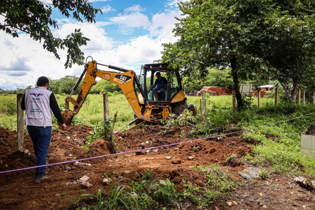 En un entorno rural con vegetación y árboles, un profesional de espaldas lleva un chaleco blanco institucional con la inscripción "UBPD - Misión Humanitaria". A su derecha, una retroexcavadora Caterpillar color amarillo remueve tierra en una zona delimitada por un cerramiento sencillo.