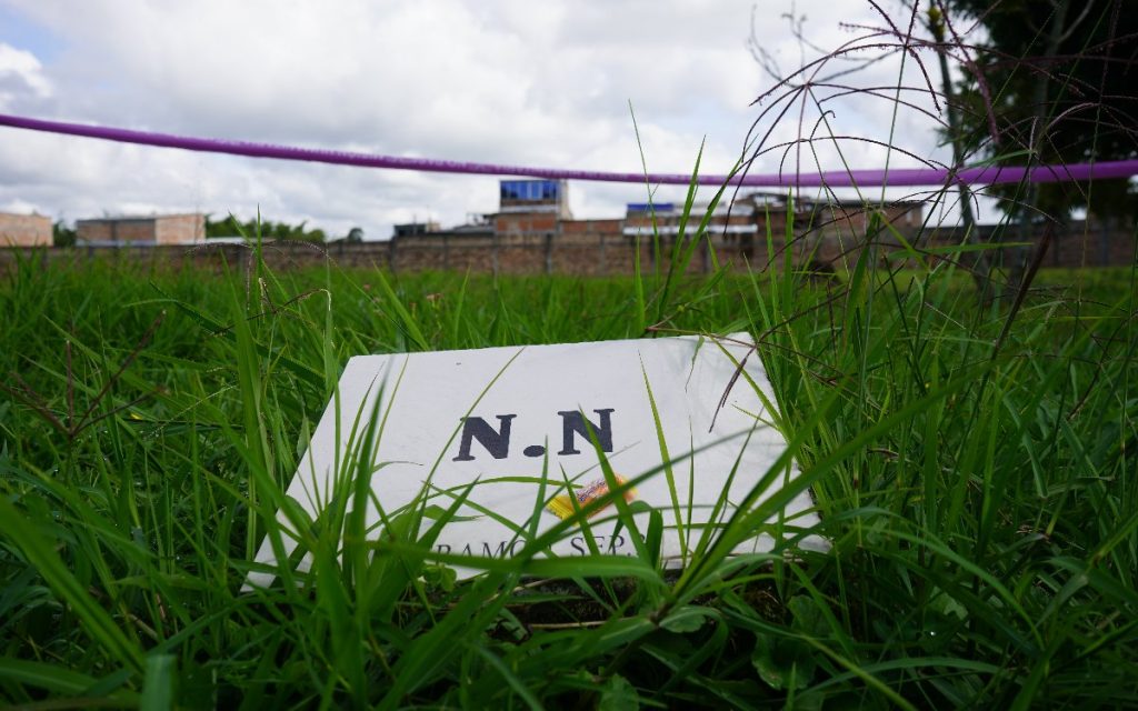 Toma en primer plano desde el nivel del pasto donde se observa una placa de piedra o cemento con las iniciales "N.N.". Sobre la placa hay un pequeño dulce empaquetado. Al fondo, tras una hilera de hierba y delimitado por una cinta de seguridad morada, se observan construcciones del entorno urbano.