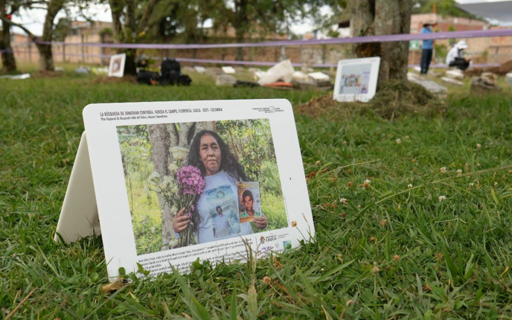 Primer plano de una pancarta informativa en forma de caballete colocada sobre el pasto. En ella se aprecia la imagen de una mujer madura sosteniendo un ramo de flores y la fotografía impresas de un joven desaparecido. Al fondo, desenfocados, se observan árboles y personas trabajando en un espacio delimitado con cinta de seguridad.