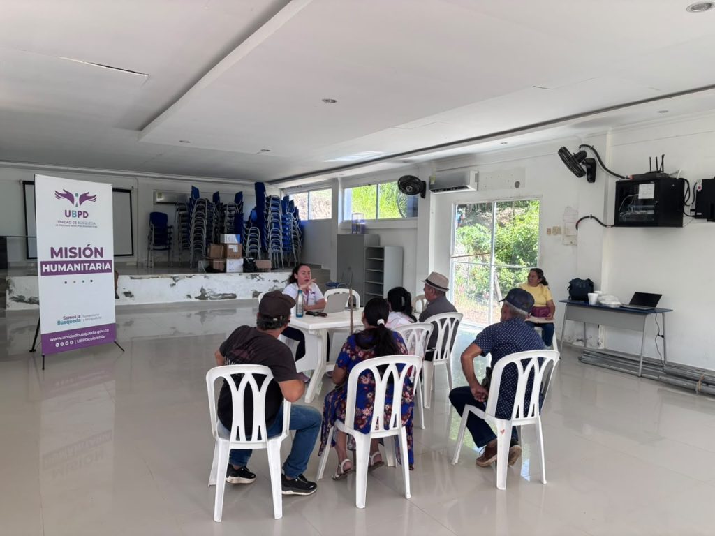 Fotografía de una reunión comunitaria en un salón claro. Siete personas están sentadas en sillas plásticas blancas alrededor de una mesa de plástico, escuchando a una funcionaria que tiene una computadora portátil. Al lado izquierdo del recinto destaca un pendón publicitario con el logotipo de la UBPD y la frase "Misión Humanitaria".