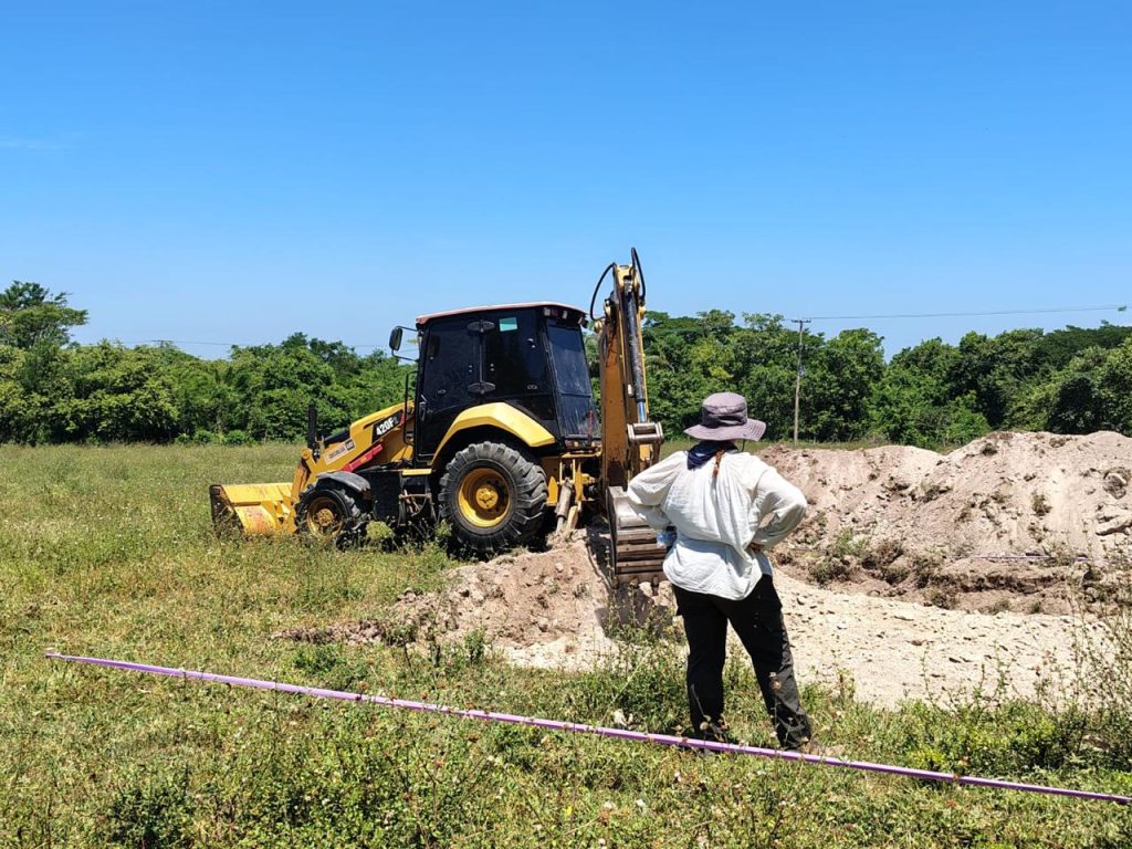 Una máquina retroexcavadora de color amarillo remueve tierra en un área rural bajo un día soleado. En primer plano, una persona vestida con ropa de trabajo de campo, sombrero de ala ancha y las manos en la cintura, observa la zanja de tierra extraída. Delante se observa una cinta delimitadora de color morado.