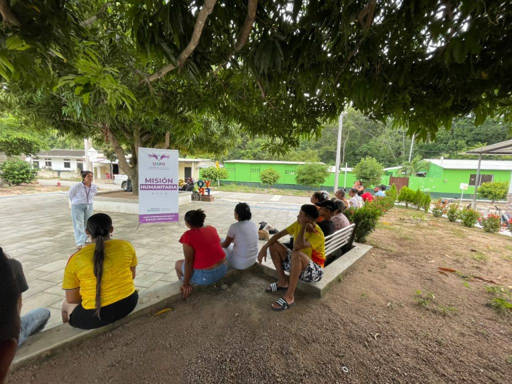 Varias personas pertenecientes a la comunidad permanecen sentadas en el borde de una jardinera y en bancas, a la sombra de un gran árbol. Frente a ellas, una funcionaria de la UBPD está de pie junto a un pendón vertical que dice "UBPD Misión Humanitaria", brindando una charla al aire libre. Al fondo se aprecian viviendas de color verde.