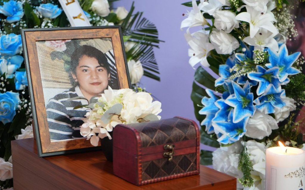 Plano medio del altar donde resalta la fotografía enmarcada de Anyi Vanessa Bravo, acompañada de un pequeño cofre de madera ornamentado, arreglos de flores blancas y lirios azules, junto a una vela encendida.