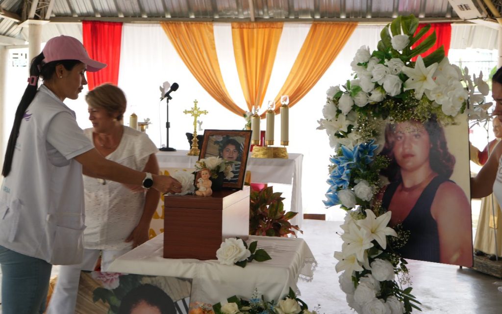 Una profesional del equipo de apoyo coloca una flor blanca sobre la urna funeraria colocada en un altar. Al lado, familiares contemplan las fotografías conmemorativas en un recinto decorado con cortinas cálidas y arreglos florales.