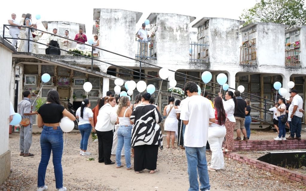 Una profesional del equipo de apoyo coloca una flor blanca sobre la urna funeraria colocada en un altar. Al lado, familiares contemplan las fotografías conmemorativas en un recinto decorado con cortinas cálidas y arreglos florales.