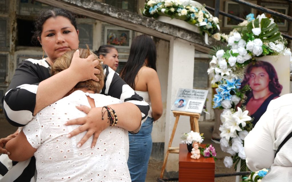 Una profesional del equipo de apoyo coloca una flor blanca sobre la urna funeraria colocada en un altar. Al lado, familiares contemplan las fotografías conmemorativas en un recinto decorado con cortinas cálidas y arreglos florales.