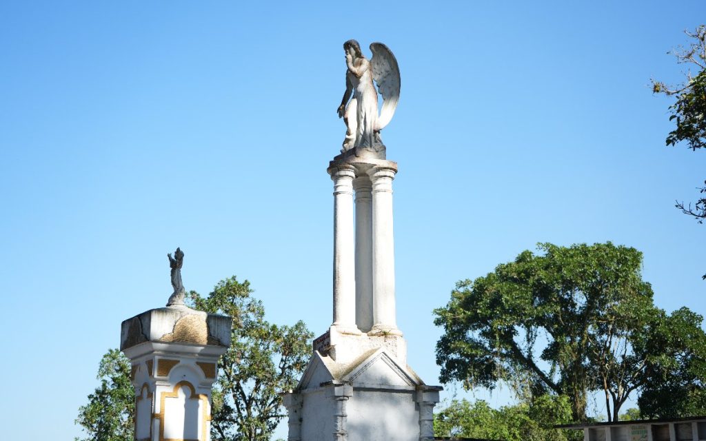 Escultura blanca de un ángel con las alas desplegadas sobre una columna alta en el Cementerio San Antonio de Padua, destacada contra un cielo azul claro y rodeada de vegetación.
