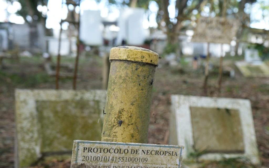 Un tubo cilíndrico amarillo marcado en una tumba con una etiqueta que dice "PROTOCOLO DE NECROPSIA 2010010141551000109", rodeado de lápidas en un cementerio.