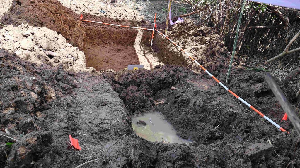 Toma cenital y de cerca que muestra un pozo excavado en la tierra con agua estancada en el fondo. Al lado se aprecia una trinchera rectangular excavada de tierra arcillosa marrón, delimitada superiormente por jalones métricos con franjas rojas y blancas y pequeñas banderas de señalización.