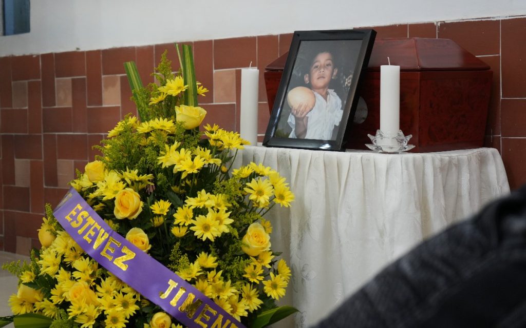 Arreglo de flores amarillas con una cinta morada que dice "ESTEVEZ JIMENEZ", junto a una mesa ceremonial con velones, el retrato de un niño y un pequeño cofre fúnebre de madera.