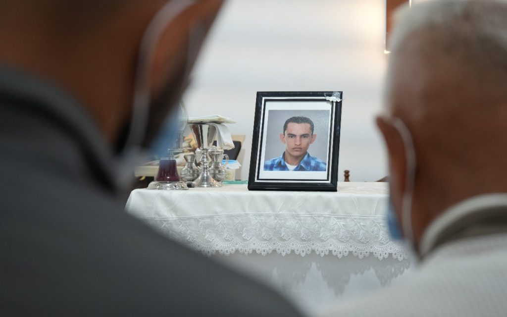 Arreglo de flores amarillas con una cinta morada que dice "ESTEVEZ JIMENEZ", junto a una mesa ceremonial con velones, el retrato de un niño y un pequeño cofre fúnebre de madera.