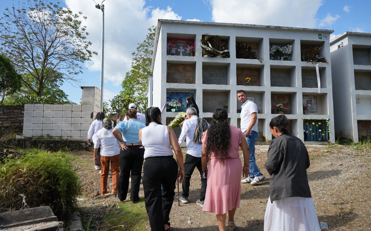 Un grupo de personas camina de espaldas frente a una estructura blanca de tumbas o nichos funerarios en un cementerio al aire libre en un día soleado.