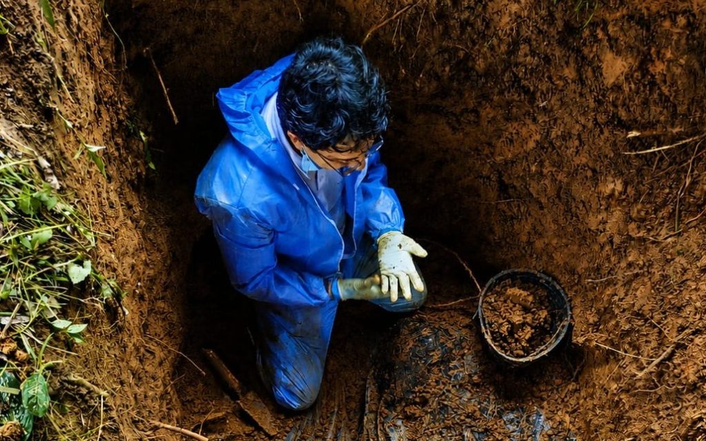 Vista desde arriba de una persona con traje de protección azul, mascarilla y guantes, arrodillada dentro de una excavación profunda en la tierra junto a un balde con lodo.