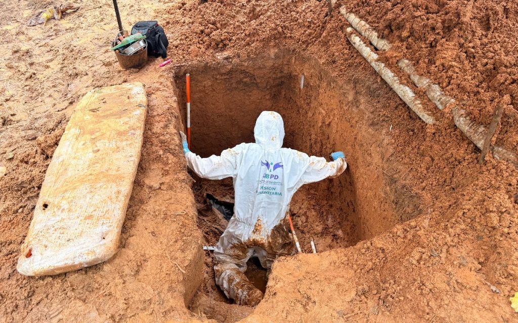 Una persona con traje de protección blanco cubierto de lodo, con el logotipo de la UBPD Misión Humanitaria, trabaja dentro de una fosa rectangular excavada en la tierra.
