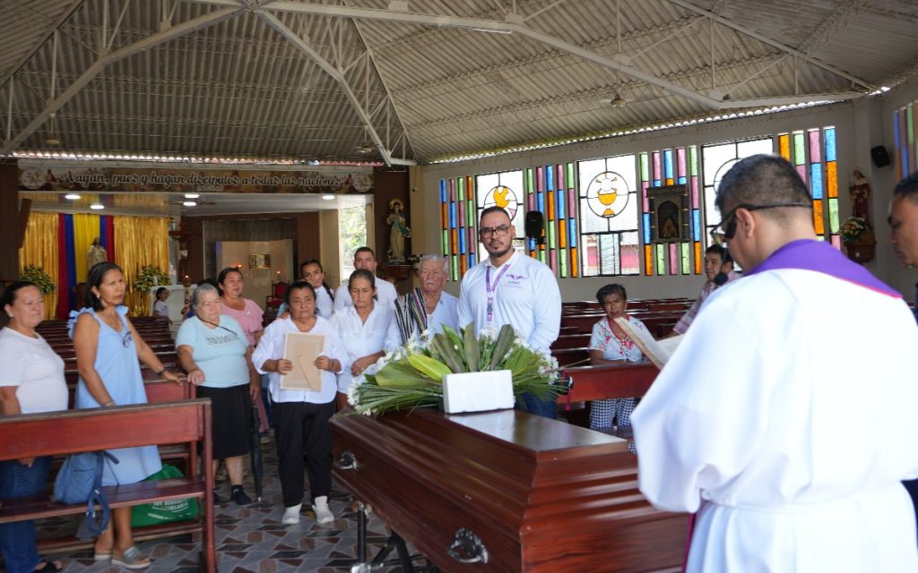 Fotografía en el interior de una iglesia iluminada por ventanales de vitrales de colores. En primer plano, de espaldas, se observa a un sacerdote con túnica blanca y estola morada sosteniendo un libro. Frente a él hay un ataúd de madera color marrón con un arreglo floral encima. Detrás del féretro se encuentran reunidas varias personas, entre ellas familiares con vestimentas sencillas y un funcionario de la Unidad de Búsqueda de Personas dadas por Desaparecidas (UBPD) con camisa blanca institucional y gafas.