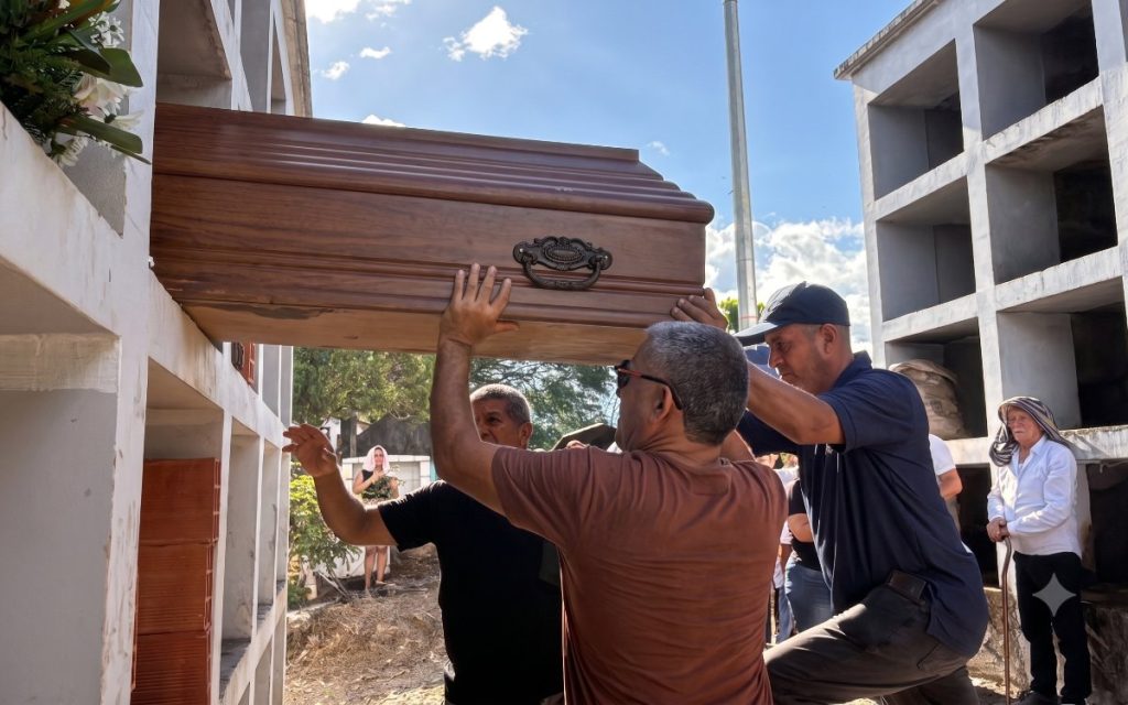 Plano medio en ángulo contrapicado que muestra a tres hombres con esfuerzo físico levantando en alto un féretro de madera color marrón claro para ingresarlo en uno de los espacios elevados de una pared de bóvedas de concreto. Al fondo se distingue a un adulto mayor observando la escena.