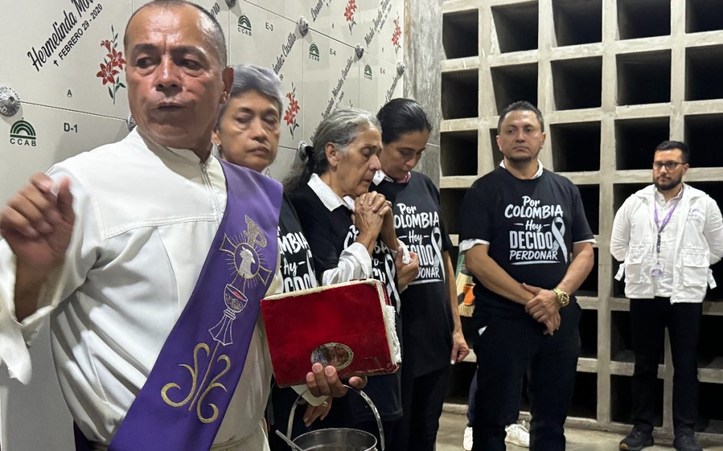 Un sacerdote católico con alba blanca y estola morada sostiene un libro litúrgico rojo y un recipiente con agua bendita frente a un grupo de personas en un área de osarios de un cementerio. Una mujer anciana reza con las manos juntas, mientras otros familiares escuchan solemnemente portando camisetas negras con la frase impresa "Por Colombia hoy decido perdonar".