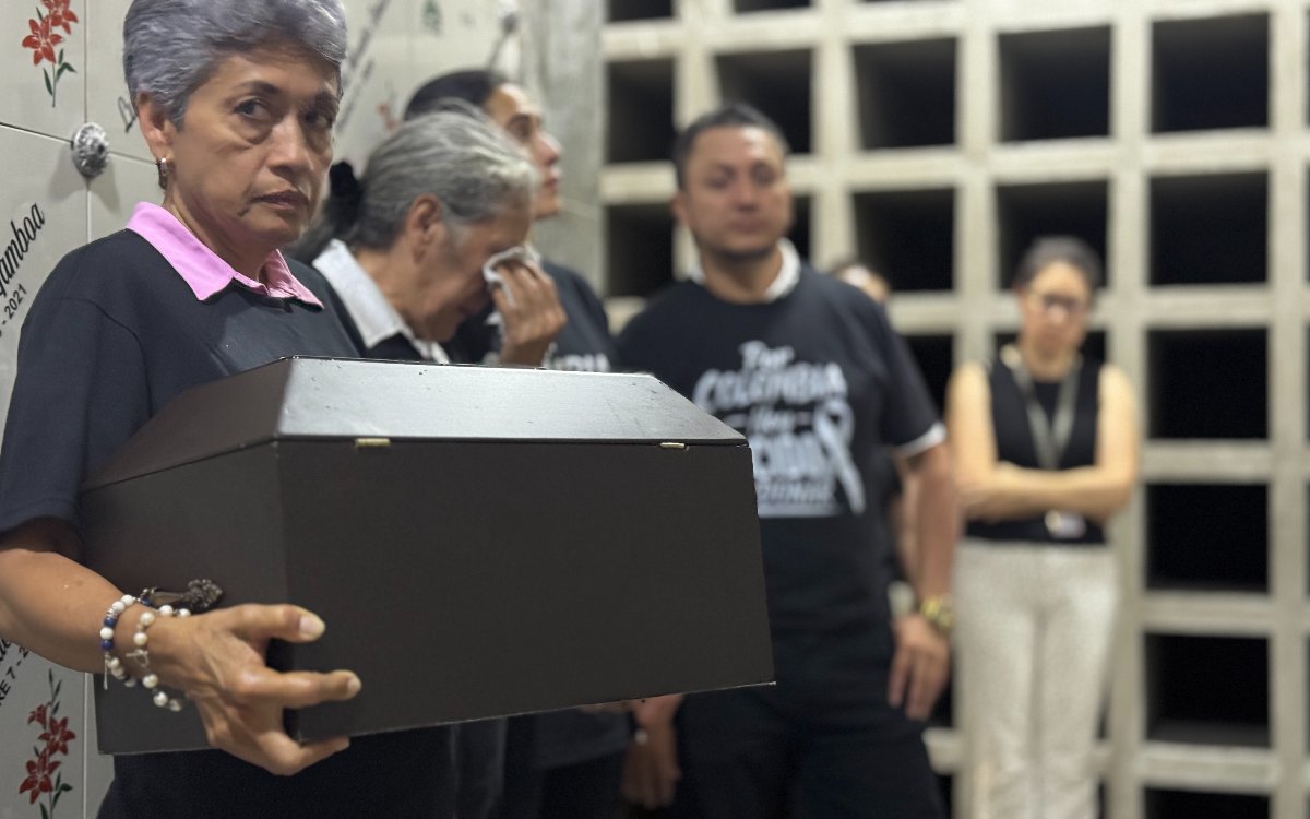 Plano medio de una mujer de cabello corto y canoso que sostiene con firmeza una pequeña urna o cofre funerario de madera. Detrás de ella, otra mujer se limpia las lágrimas y un hombre con una camiseta negra con el mensaje 