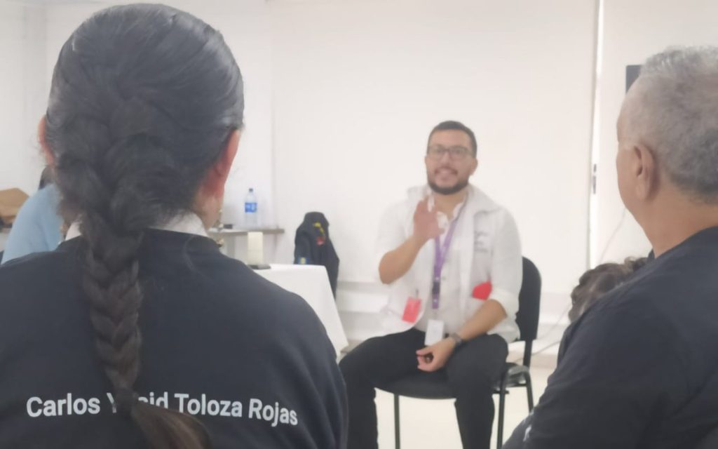 Vista desde la espalda de una persona con el cabello recogido en una trenza, vistiendo una camiseta negra con el nombre impreso "Carlos Yair Toloza Rojas". En frente, en un plano desenfocado, un hombre con gafas y chaleco institucional habla gestualizando con las manos durante un taller o espacio de diálogo.