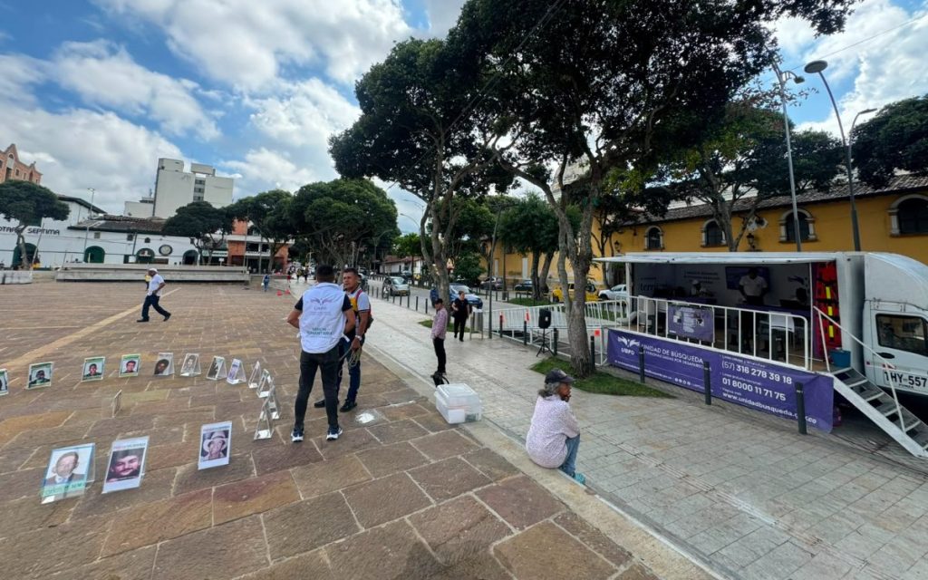 Fotografía de una plaza adoquinada al aire libre donde se exhiben varios retratos enmarcados de personas desaparecidas colocados en el suelo. A la derecha, se encuentra estacionada una unidad móvil de atención con pancartas e información de contacto de la Unidad de Búsqueda de Personas dadas por Desaparecidas (UBPD) de Colombia. Un funcionario con chaleco de la UBPD dialoga con un ciudadano en la plaza.