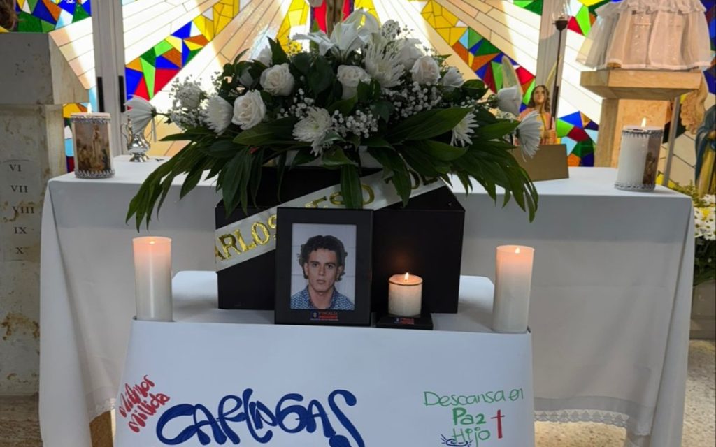 Altarcito funerario sobre una mesa cubierta con un mantel blanco frente a un ventanal de vitrales coloridos. Al centro hay un cofre o urna rodeado por un gran arreglo floral de lirios y rosas blancas, una cinta con el nombre "Carlos" y un retrato enmarcado de un joven. A los lados hay velas blancas encendidas y en la parte inferior se leen mensajes escritos a mano como "Carlangas", "Mi amor milida" y "Descansa en Paz Hijo".
