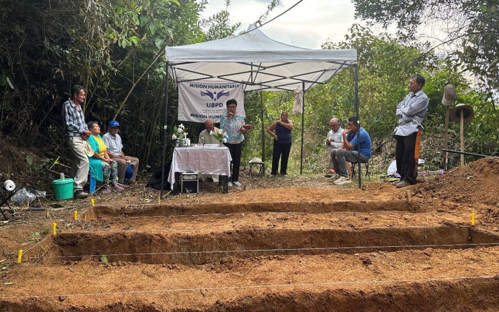 Fotografía en zona rural donde se observa una carpa blanca institucional con un cartel al fondo que dice «MISIÓN HUMANITARIA - UBPD». Alrededor de una mesa y bajo la carpa hay varias personas sentadas y de pie escuchando a una mujer que habla por un micrófono. En primer plano se aprecian zanjas rectangulares delimitadas por hilos en el suelo de tierra.