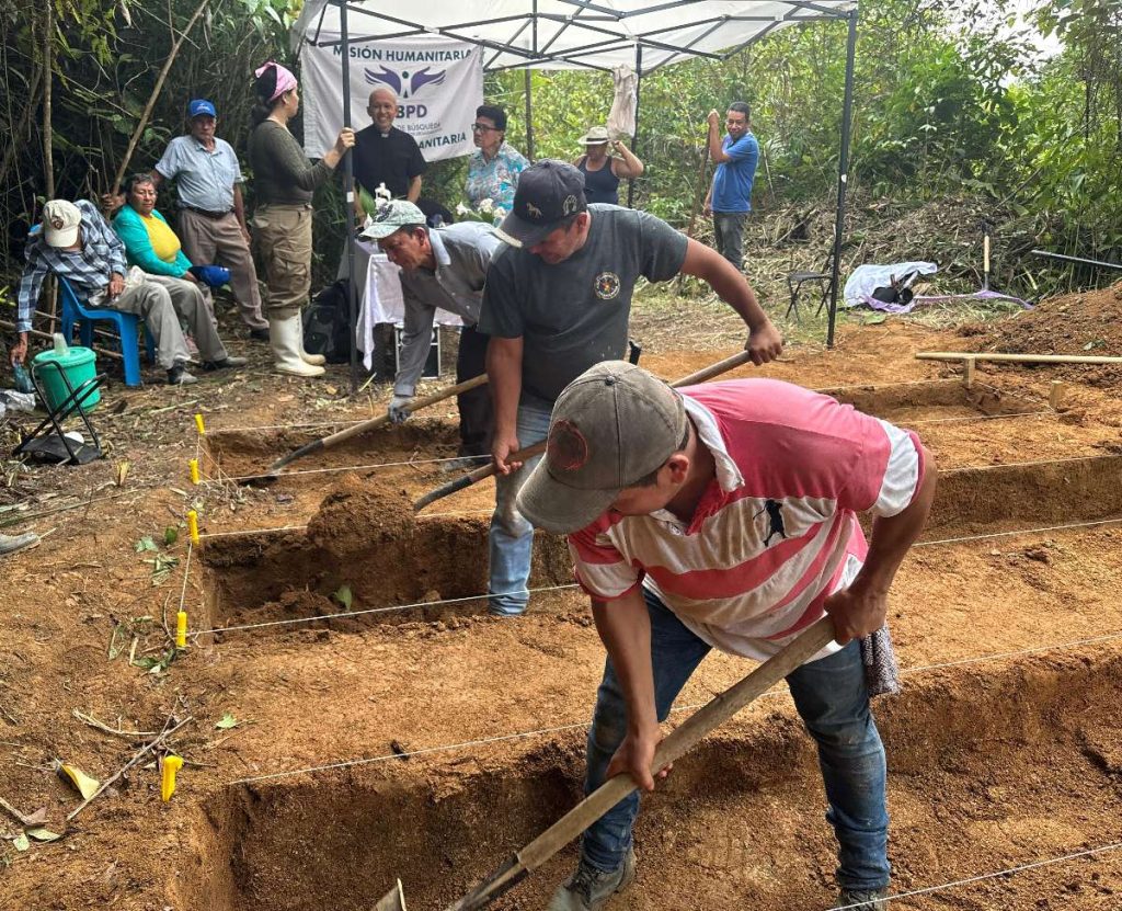 En primer plano, un hombre con camiseta a franjas rosa y blanca y gorra utiliza una pala para cavar en una zanja de tierra. A su lado, otro campesino colabora en la excavación. En el fondo se ve a miembros del equipo humanitario y personas de la comunidad observando el proceso bajo una carpa.