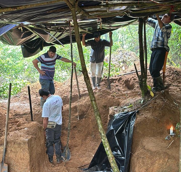 Varios hombres trabajan dentro de un terreno excavado de ladera cubierto por una estructura provisional de madera y plástico negro para proteger el área de la lluvia. En el costado derecho del terreno hay una vela blanca encendida colocada sobre un altar improvisado.