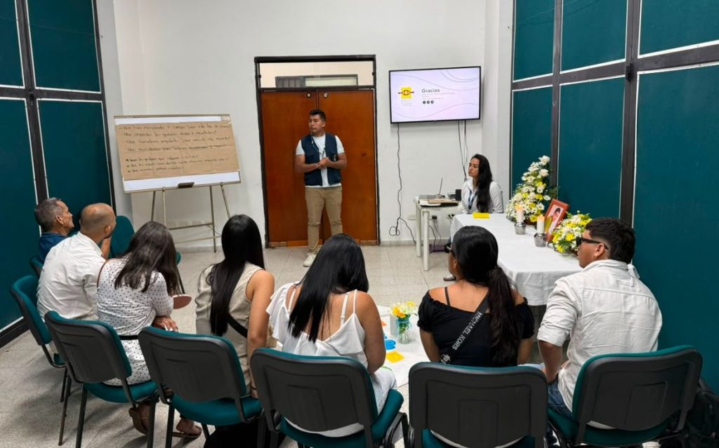 Un grupo de personas sentadas en semicírculo escucha la presentación de un funcionario de la UBPD frente a una mesa ceremonial con flores y una fotografía.
