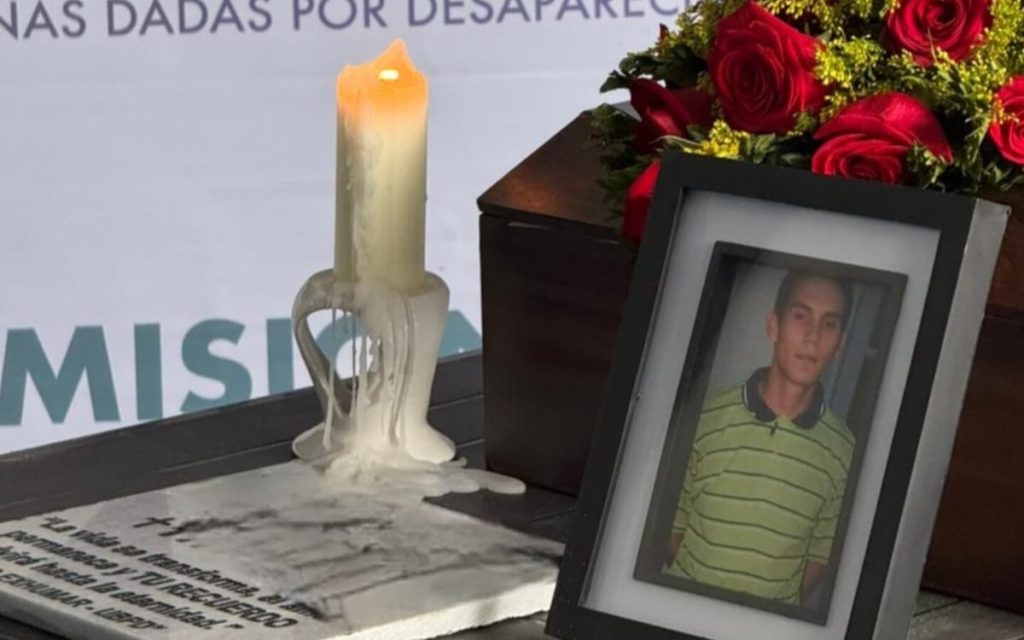 Primer plano de un retrato enmarcado de un hombre joven junto a una vela encendida con cera derretida y arreglos de rosas rojas sobre una mesa ceremonial.