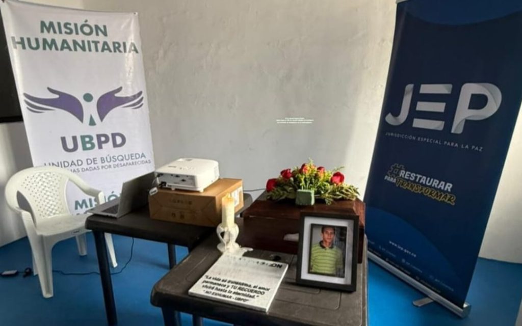 Altas con logos de la UBPD y la JEP rodean una mesa de apoyo y una mesa ceremonial con el cofre funerario, la foto de un joven y flores.
