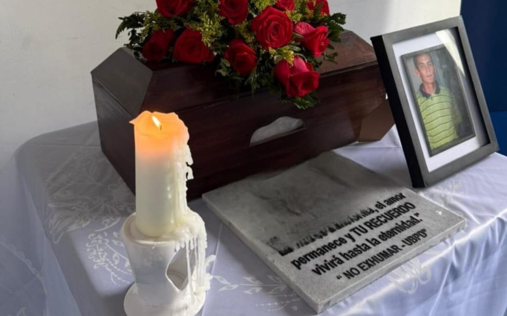 Mesa altar cubierta con un cofre de madera, un arreglo de rosas rojas, una velón encendido, un retrato masculino y una placa con la frase: "La vida se transforma, el amor permanece y TU RECUERDO vivirá hasta la eternidad. NO EXHUMAR - UBPD".