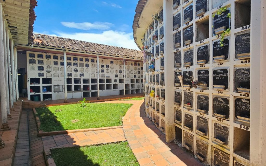 Perspectiva de un patio en el cementerio de Granada. A la derecha se aprecia una estructura semicircular con nichos numerados e identificados con placas de mármol negro y arreglos florales. Un pasillo con baldosas cerámicas terracota rodea las áreas verdes centrales, cubierto por un tejado de tejas de arcilla sostenido por columnas blancas.